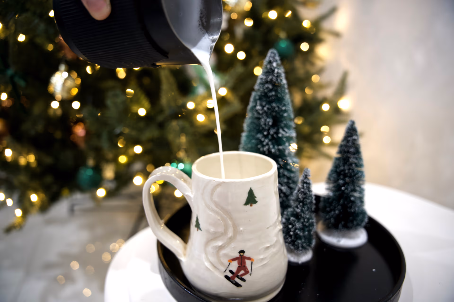 Milk being poured into a wintery mug in front of a tree with lights on it.
