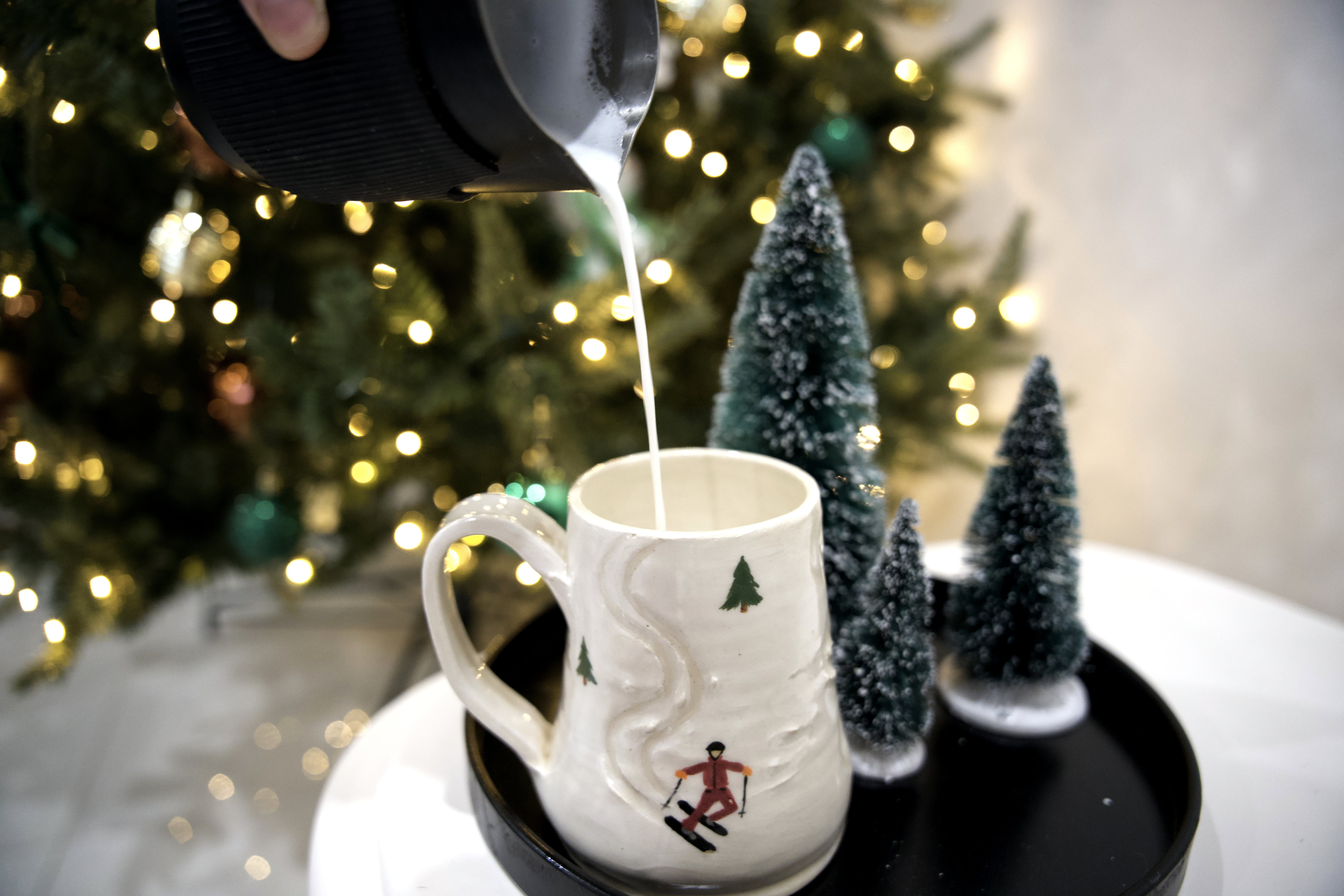 Milk being poured into a wintery mug in front of a tree with lights on it. 
